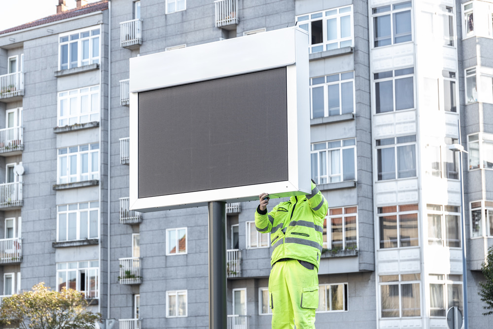 A worker wearing reflective safety gear installs a rectangular LED display sign in front of an apartment building.